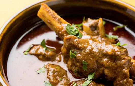 A bowl of rich paya curry with tender goat trotters in a deep red masala gravy, garnished with fresh coriander, green chillies, and a wedge of lemon, served alongside naan bread.