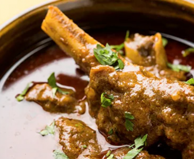 A bowl of rich paya curry with tender goat trotters in a deep red masala gravy, garnished with fresh coriander, green chillies, and a wedge of lemon, served alongside naan bread.