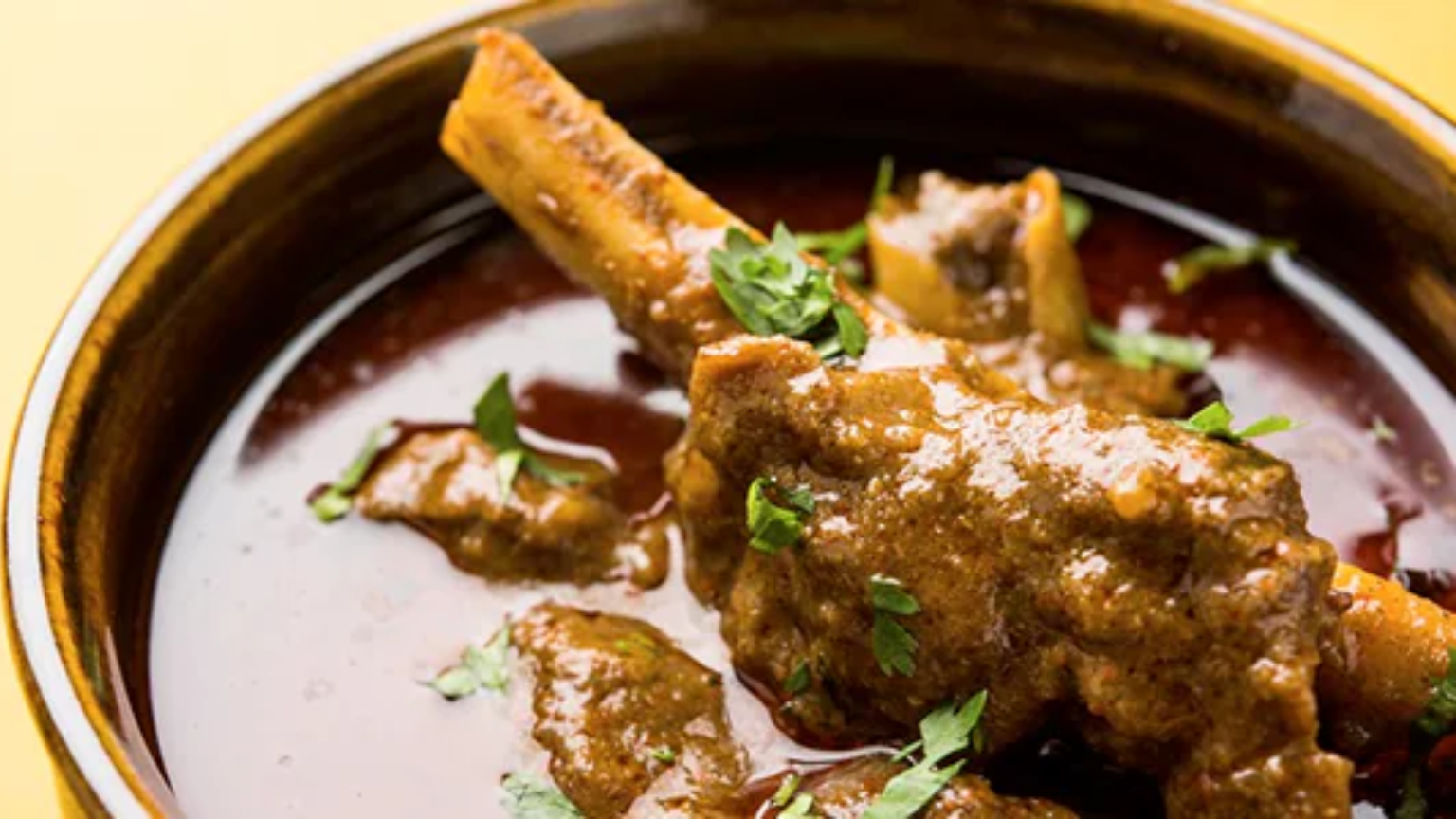 A bowl of rich paya curry with tender goat trotters in a deep red masala gravy, garnished with fresh coriander, green chillies, and a wedge of lemon, served alongside naan bread.